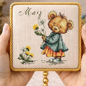 Op de afbeelding: Kruissteekkunstwerk met een teddybeer die paardenbloemen vasthoudt, gekleed in een teal trui en oranje rok. Het woord "May" is bovenaan gestikt. Het kunstwerk is ingelijst in een vierkant met een gouden touwrand en een gouden kwast.