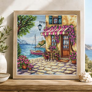 Puede incluir: Un punto de cruz enmarcado que representa una escena costera. La obra de arte presenta un edificio con un toldo a rayas, un velero y flores exuberantes. La paleta de colores incluye azules, rosas, verdes y marrones, creando un paisaje encantador y detallado.