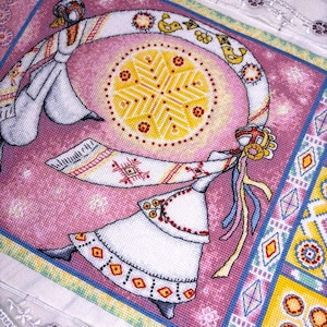 May include: A cross-stitch textile featuring two figures in white traditional clothing, dancing around a central yellow sunburst design. The background is pink with geometric patterns in blue, yellow, and red.