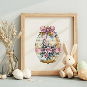 Puede incluir: Un diseño de punto de cruz enmarcado de un huevo de Pascua decorado con un lazo rosa y detalles florales. El huevo es blanco con detalles dorados y una cinta rosa.