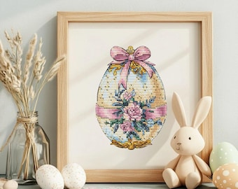 May include: A framed cross-stitch design of a decorated Easter egg with a pink bow and floral accents. The egg is white with gold accents and a pink ribbon.