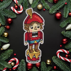 May include: A cross-stitched Christmas ornament of a nutcracker soldier in a red uniform and hat, standing on a drum. The ornament is surrounded by Christmas decorations, including candy canes, ornaments, and evergreen branches.