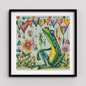 Puede incluir: Arte de punto de cruz de una rana verde con un diseño caprichoso. La rana está rodeada de corazones, flores y setas de colores. La obra de arte está enmarcada en un marco cuadrado negro.