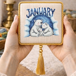 Puede incluir: Un bordado de punto de cruz enmarcado que representa una familia de osos polares en una cueva de hielo azul. La palabra "JANUARY" está bordada en la parte superior. El marco es beige con un borde de cuerda dorada y una borla dorada.