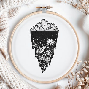 May include: A circular wooden embroidery hoop containing white fabric with a black cross-stitch design. The pattern features a mountain range at the top, transitioning into a deep space scene below, filled with various planets, stars, and rings.