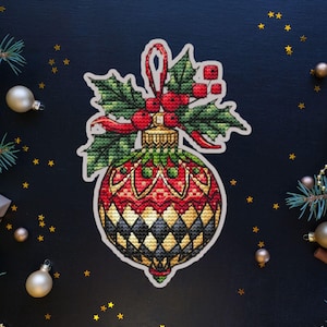 Puede incluir: Un adorno navideño rojo, dorado y verde con una rama de acebo y bayas rojas. El adorno tiene un patrón de diamantes y está decorado con detalles dorados.