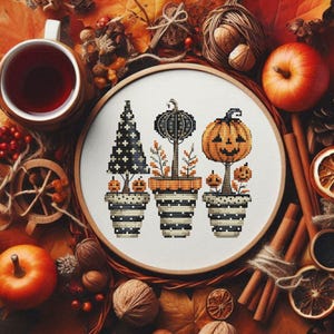May include: A cross-stitch embroidery hoop featuring three potted trees with pumpkin and Halloween designs. The trees are black, orange, and white, set against a backdrop of fall leaves, apples, and cinnamon sticks.