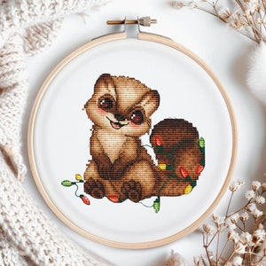 May include: Cross-stitch embroidery of a cartoon otter with brown fur, large eyes, and a smiling expression. The otter is wrapped in a string of colorful Christmas lights. The embroidery is set in a wooden hoop.