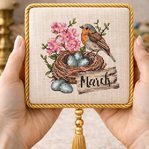 May include: A square, framed cross-stitch featuring a bird perched on a nest with eggs, surrounded by pink flowers and green leaves. The word "March" is stitched on a wooden sign. The frame has a gold rope trim and a gold tassel.