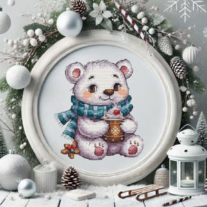 May include: A framed cross-stitch of a polar bear wearing a teal scarf, holding a cup of whipped cream and a gingerbread cookie. The bear is surrounded by a white frame and winter decorations, including ornaments and pine cones.