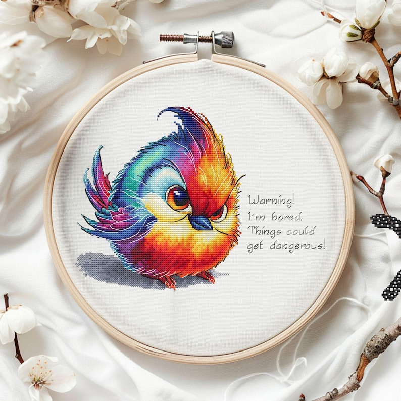 May include: A cross-stitch embroidery hoop with a colorful bird design. The bird has a red, orange, yellow, blue, and purple plumage. The bird is looking at the viewer with an angry expression. The text "Warning! I'm bored. Things could get dangerous!" is written below the bird.