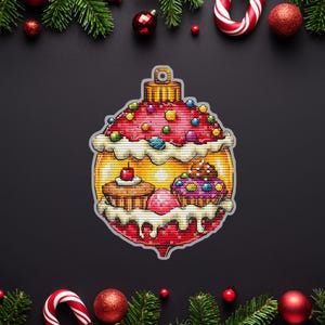 May include: A cross-stitch Christmas bauble ornament with a cupcake design. The ornament is mainly red with yellow and white details, decorated with colourful beads and frosting. The background is black with festive decorations.