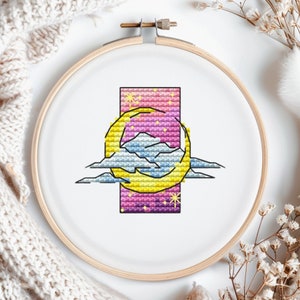 Könnte beinhalten: Ein Kreuzstich-Stickrahmen mit einem Himmelsdesign. Das Kunstwerk zeigt einen Halbmond, Wolken und Sterne vor einem farbverlaufenden Hintergrund aus Rosa, Lila und Gelb. Die Stickerei befindet sich in einem Holzrahmen.
