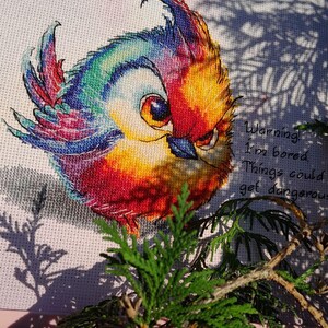 May include: A colorful cross-stitch embroidery of a bird with the text "Warning! I'm bored. Things could get dangerous."