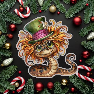May include: A cross-stitch design of a cartoon snake wearing a green and pink top hat with orange hair. The snake has large green eyes and a smiling expression. The design is surrounded by Christmas decorations, including ornaments, candy canes, and pine branches.