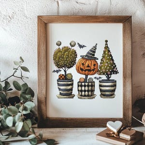 May include: A framed cross-stitch embroidery featuring three potted plants with Halloween themes: a round green topiary, a central orange jack-o'-lantern wearing a striped witch hat, and a triangular green tree, all accented by small flying bats.