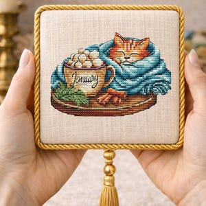 Op de afbeelding: Kruissteekkunst met een slapende oranje kat die in een blauwe deken is gewikkeld. Een mok warme chocolademelk met marshmallows en het woord "January" zijn ook afgebeeld. Het kunstwerk is omlijst met een gouden touwrand en een kwast.