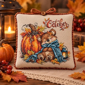 Peut inclure: Un coussin carré en tissu avec une broderie au point de croix représentant un ours en peluche marron enveloppé dans une écharpe bleue, assis près d'une grosse citrouille orange et d'une lanterne. Le mot October est brodé en rouge.
