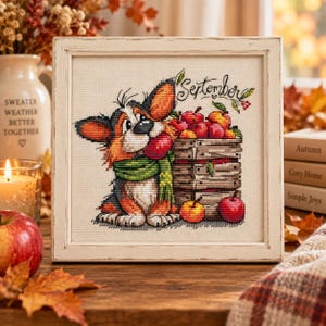 May include: A framed cross-stitch embroidery featuring a tricolor dog wearing a green scarf, holding a red apple in its mouth next to a wooden crate filled with red and yellow apples. The text September is stitched in black above the crate.