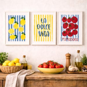 Könnte beinhalten: Drei gerahmte Kunstdrucke mit weißen Rahmen. Einer zeigt Zitronen auf blau-weißen Streifen, ein anderer liest "LA DOLCE VITA" auf gelben Streifen, und der dritte zeigt Tomaten auf blauen Streifen mit dem Wort "POMODORO". Ein rustikaler Holztisch zeigt Zitronen, Tomaten und Küchenartikel.