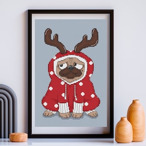 May include: A framed cross-stitch pattern featuring a tan pug dog wearing a red hooded sweatshirt with white polka dots and brown reindeer antlers. The pug has a black muzzle and is depicted against a light blue background with a grid texture.