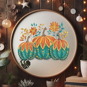 May include: A cross-stitch embroidery featuring three pumpkins in teal, orange, and yellow. The pumpkins are adorned with floral and leaf accents, set against a cream-colored background within a round wooden frame. The artwork evokes a fall harvest theme.