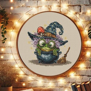 May include: A cross-stitch embroidery of a cartoon frog wearing a witch's hat and sitting in a cauldron. The frog is green with large eyes, and the hat is blue with star details. A small broom is next to the cauldron.