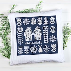 Op de afbeelding: Wit vierkant kussen met een marineblauw kruissteekontwerp. Het ontwerp bevat bloemen, huizen en geometrische patronen, waaronder bloemen, sneeuwvlokken, een uil en een peer. Het kussen ligt op een wit oppervlak met groen gebladerte.
