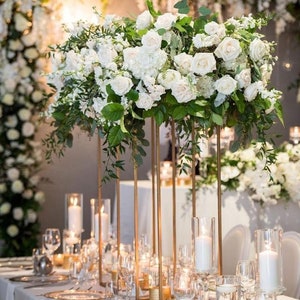 Op de afbeelding: Een bruiloft centerpiece met witte rozen en groen, gerangschikt in een hoge gouden metalen standaard. Het centerpiece is omringd door kaarsen en heldere glazen vazen op een wit tafelkleed.