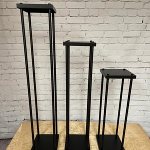 Peut inclure: Trois supports à fleurs en métal noir de hauteurs différentes. Les supports sont faits de tubes carrés et ont une base carrée.