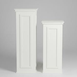 May include: Two white rectangular pedestals of different heights, each featuring a flat top surface and a recessed panel design on the front face. The structures are constructed with clean, straight lines and a smooth, solid white finish.