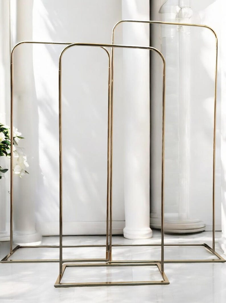 May include: Three gold metal arch stands with square bases. The stands are tall and slender, and they are perfect for displaying flowers or other decorations.