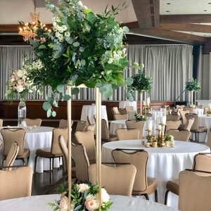 May include: A tall gold metal stand with a square base holds a large arrangement of greenery and white flowers. The stand is placed on a white tablecloth on a round table in a room with beige chairs and white tablecloths.