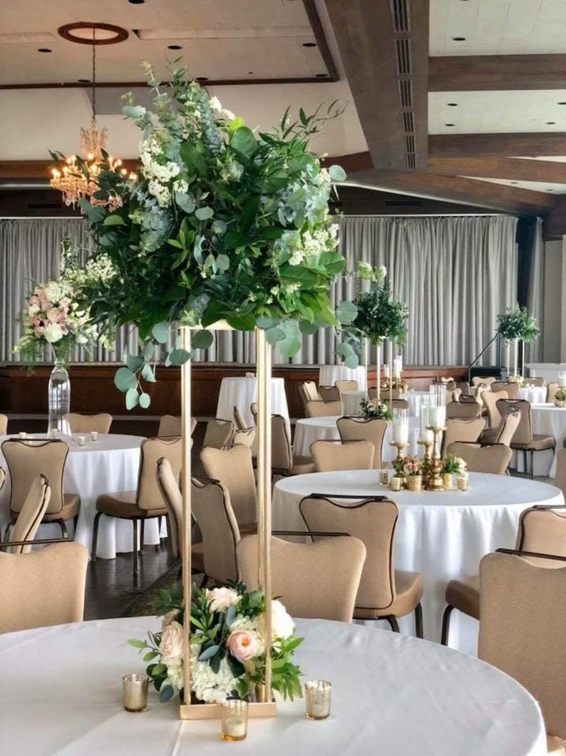 May include: A tall gold metal stand with a square base holds a large arrangement of greenery and white flowers. The stand is placed on a white tablecloth on a round table in a room with beige chairs and white tablecloths.