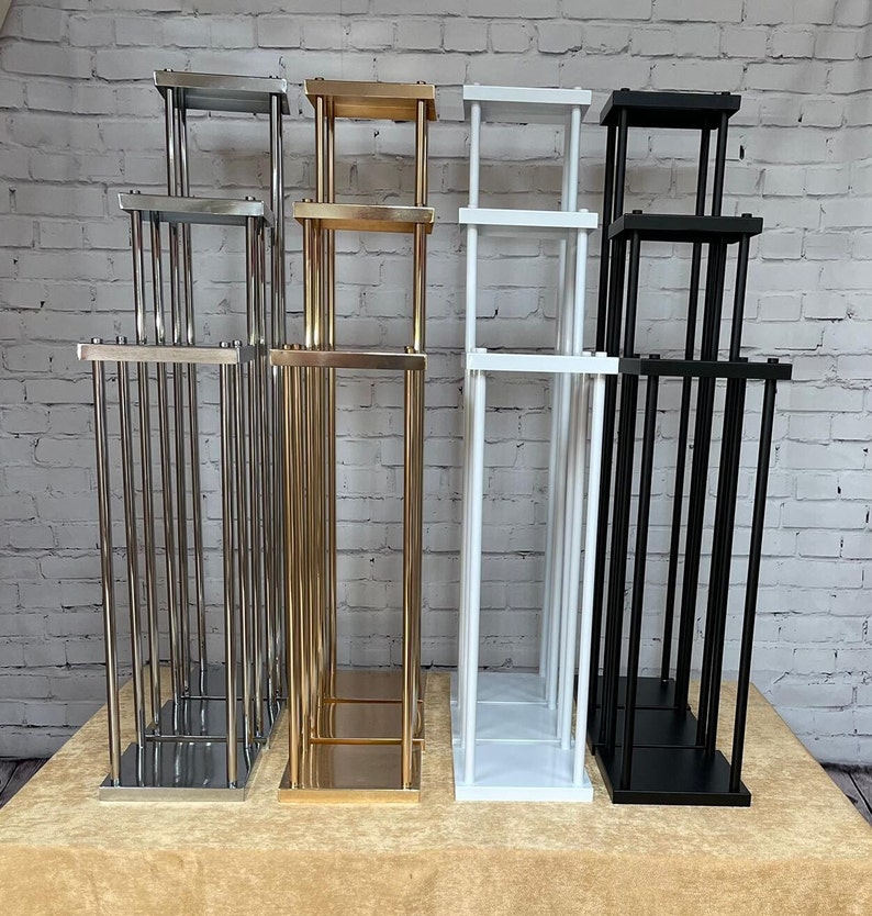 May include: Four metal flower stands in different colors: silver, gold, white, and black. Each stand has three tiers and is made of thin metal rods.