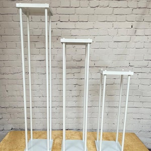 May include: Three white metal flower stands of varying heights. The stands are made of thin metal rods and have square bases.