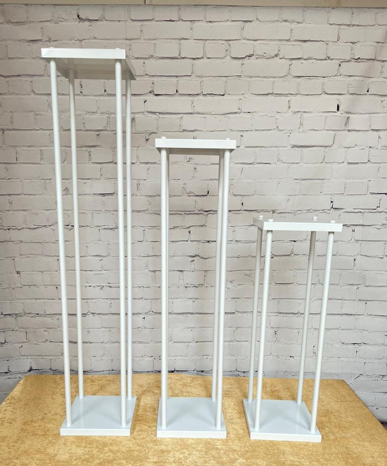May include: Three white metal flower stands of varying heights. The stands are made of thin metal rods and have square bases.