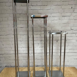 May include: Three silver metal flower stands of varying heights. The stands are made of thin metal tubing and have square bases.