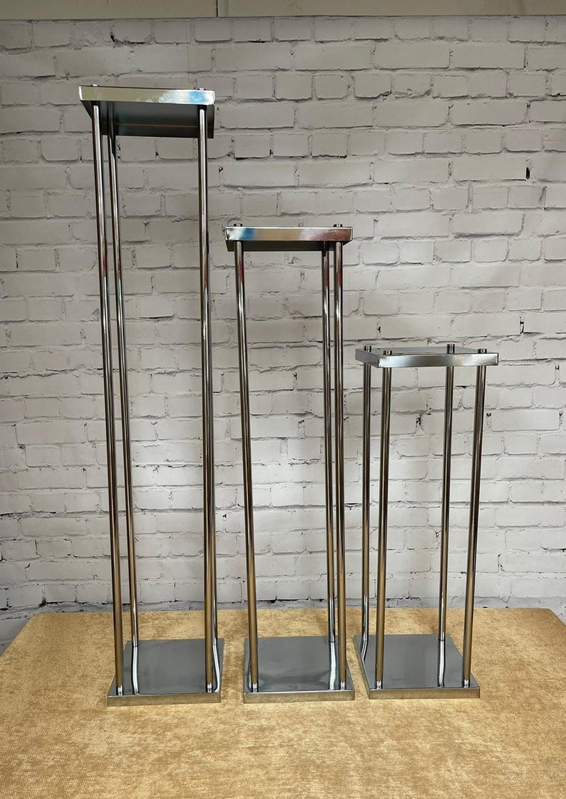 May include: Three silver metal flower stands of varying heights. The stands are made of thin metal tubing and have square bases.