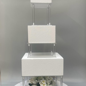 Op de afbeelding: Een drielaags witte taartstandaard met transparante acryllagen en een witte marmeren basis. De standaard is versierd met een wit bloemstuk.