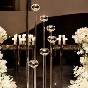 Può includere: Un set di portacandele alti, color oro, con sommità sferiche. I supporti sono disposti verticalmente, con altezze variabili. Lo sfondo presenta candele e composizioni floreali, creando un'atmosfera elegante.