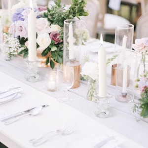 Può includere: Un'elegante tavola imbandita con tovaglie bianche, posate e composizioni floreali. Candele accese in portacandele di vetro e vasi con fiori creano un'atmosfera sofisticata. La palette di colori è delicata e invitante, perfetta per occasioni speciali.