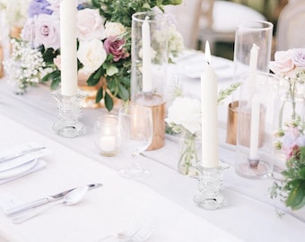 May include: A beautifully arranged table setting with white linens, silverware, and floral centerpieces. Tall, lit candles in glass holders and clear glass vases with flowers create an elegant ambiance. Wine glasses and small votive candles add to the decor.