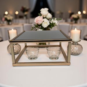 May include: A gold metal and glass decorative centerpiece with a mirrored top, two small glass bowls, and a floral arrangement. Two lit candles in glass holders are on either side. The table is white.