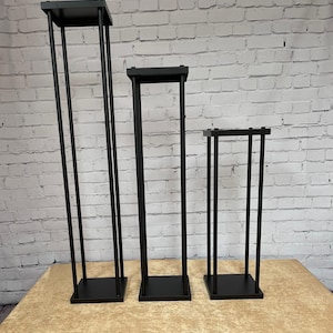 May include: Three black metal flower stands of varying heights. The stands are made of square tubing and have a square base.