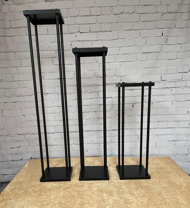 May include: Three black metal flower stands of varying heights. The stands are made of square tubing and have a square base.