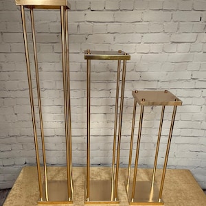 May include: Three gold metal flower stands of varying heights. The stands are square and have a simple, modern design.