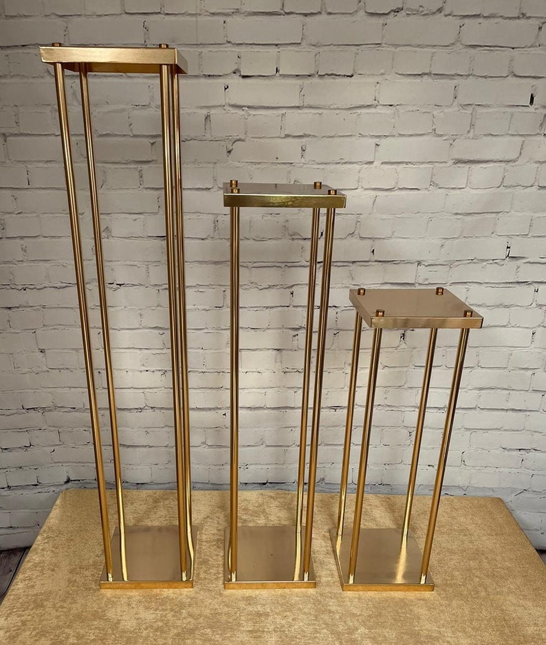 May include: Three gold metal flower stands of varying heights. The stands are square and have a simple, modern design.