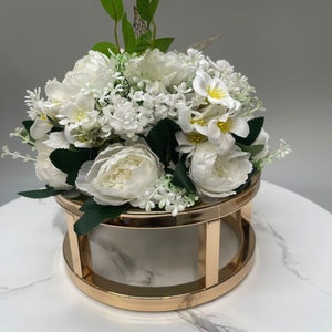 Op de afbeelding: Een wit kunstbloemenarrangement in een gouden metalen standaard. De bloemen zijn in een ronde vorm gerangschikt en omvatten witte rozen, witte anjers en witte gipskruid.
