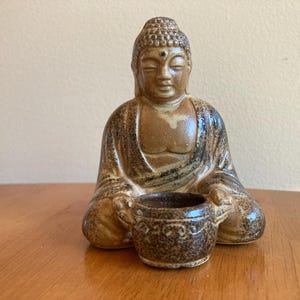 May include: A ceramic Buddha figurine in a seated meditation pose with a glazed tan and dark brown speckled finish. The figure holds a small, round, two-handled bowl in its lap, featuring a textured, dark brown surface with subtle embossed details.